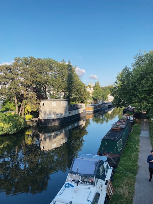A view of the Little Venice area showing narrowboats moored along a calm canal, with leafy green trees lining the waterway under a bright blue sky with some scattered clouds. The scene includes boats with polished surfaces reflecting the natural surroundings, and a pedestrian walking along the paved towpath. The lighting highlights the cleanliness and tranquility of the environment, emphasizing the importance of surface cleaning and maintenance for a hygienic, well-kept outdoor space. This image relates to the high standards of cleaning and sanitisation provided by Maida Vale Carpet Cleaning in the context of flood cleanup and deep cleaning cases in the Little Venice area.