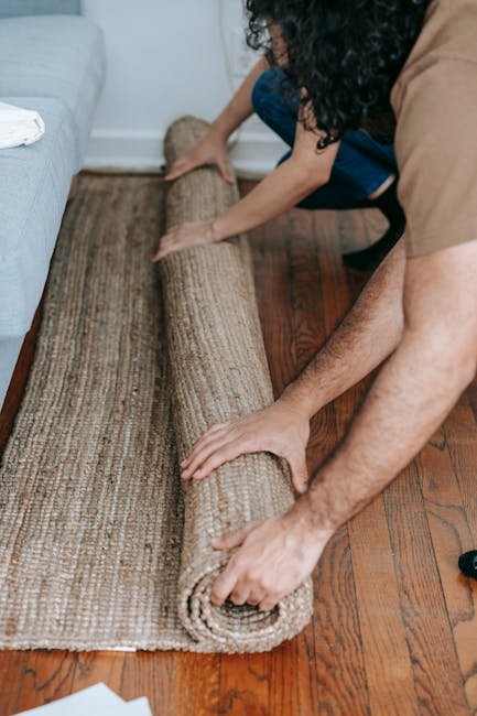 A person is carefully unrolling a beige and brown textured area rug onto a polished hardwood floor in a living room. The room features white walls and a light-colored sofa with a white cushion. The individual is wearing a brown shirt and has their hands positioned on either end of the rug, ensuring smooth placement. Visible cleaning tools or products are not present in this image. The scene highlights proper surface care and placement of rugs as part of domestic cleaning and maintenance, reflecting the expertise of Maida Vale Carpet Cleaning in deep cleaning and surface sanitisation services. Natural lighting illuminates the space, emphasizing the rug’s clean and well-maintained condition, suitable for a case study on flood cleanup and carpet cleaning in Little Venice.