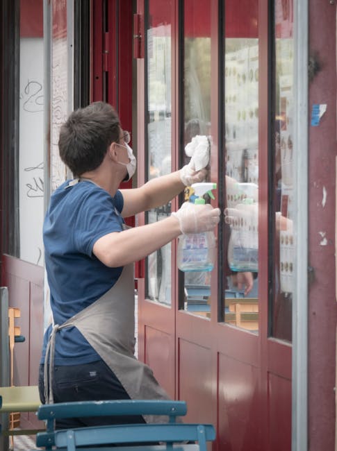 A person wearing a face mask, gloves, and a grey apron is cleaning large glass windowpanes at the entrance of a shop or café. They are using a spray bottle and a cloth to wipe the glass, which appear