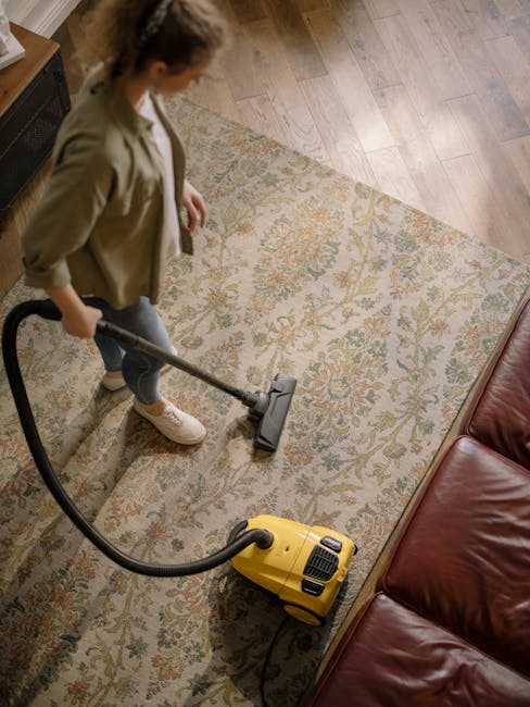 A person kneeling on a patterned carpet in a domestic setting, using a yellow wet vacuum cleaner for surface cleaning and sanitisation of the carpet. The individual is wearing a khaki jacket and blue jeans, and is adjusting the machine's hose or control panel. The room is well-lit, likely with natural light, highlighting the carpet's detailed floral design and the shiny, clean appearance after cleaning. The scene emphasizes professional carpet cleaning methods used by Maida Vale Carpet Cleaning to maintain cleanliness and hygiene, suitable for same-day service in W9 area.