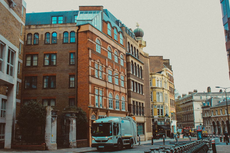 A city street scene in Maida Vale with a mix of older and modern buildings, including red brick and pastel-colored facades, large glass windows, and a decorative dome on one of the rooftops. The stree