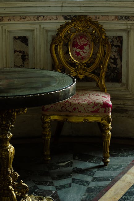 An ornate antique armchair with a gold carved wooden frame and pink floral upholstery, positioned behind a polished marble table with a dark finish. The chair is set against a decorative marble wall with intricate moldings and panels, illuminated by soft lighting that highlights the luxurious textures and surfaces. The floor features a black and white marble mosaic pattern, adding to the elegant ambiance. The scene depicts a well-maintained, historically styled interior space, emphasizing the importance of surface cleaning and preservation of period furnishings, as provided by Maida Vale Carpet Cleaning's domestic cleaning services.