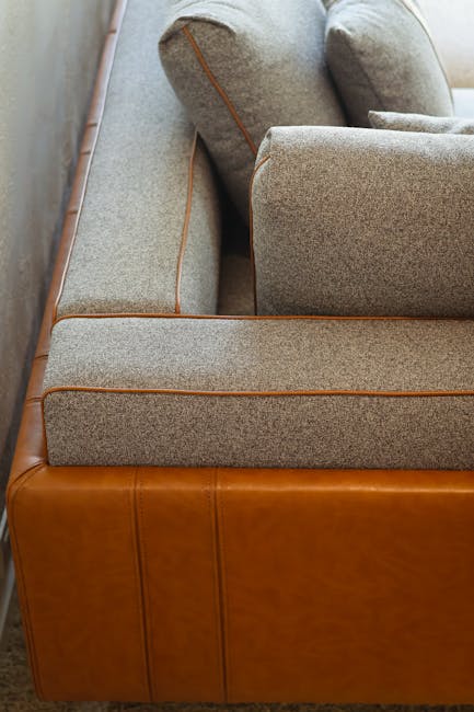Close-up view of a mid-century modern sofa with a wooden frame and light grey upholstered cushions, situated in a well-lit room. The fabric on the cushions appears clean and well-maintained, with no visible stains or dust, indicating recent surface cleaning or deep cleaning and sanitisation. The sofa's leather base has a smooth, polished appearance, contributing to its overall aesthetic and hygiene. In the background, a wooden floor is partially visible, reflecting natural light, and the arrangement suggests a comfortable, tidy living space. This scene illustrates professional domestic cleaning services provided by Maida Vale Carpet Cleaning, specializing in upholstery cleaning for period homes on Warwick Avenue.